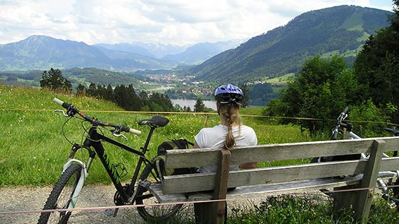 Radfahrerin macht eine kurze Rast auf einer Bank an Feldern und Wäldern